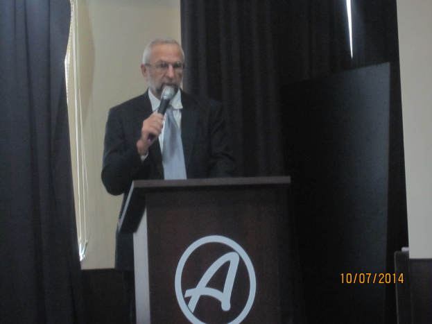 Eine Person in einem schwarzen Mantel und grauen Krawatte steht an einem Schreibtisch und hält ein Mikrofon, während sie eine Rede hält, mit schwarzen Vorhängen im Hintergrund.