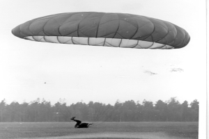Schwarzes und weißes Foto einer Person, die mit einem Gleitschirm fliegt und von einem Fallschirm umgeben ist, umgeben von Bäumen und einem klaren Himmel, mit Text unten.
