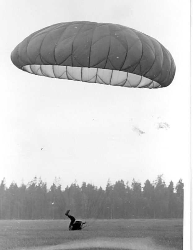 Schwarzes und weißes Foto einer Person, die mit einem Gleitschirm fliegt und von einem Fallschirm umgeben ist, umgeben von Bäumen und einem klaren Himmel, mit Text unten.