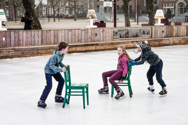 Kinder beim Skifahren vor einem Spielplatz im Vordergrund, mit drei Kindern und zwei Stühlen in der Mitte und Gebäuden, Bäumen, Bänken, Pfosten und einem Basketballfeld im Hintergrund.