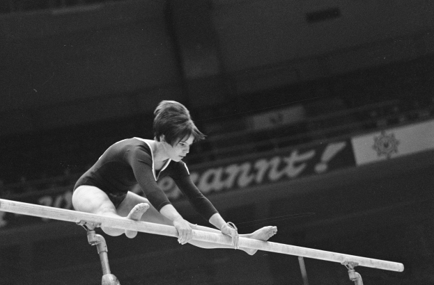Eine Frau führt Gymnastik an den parallelen Stangen in einem Schwarz-Weiß-Foto aus, mit einem Banner im Hintergrund.