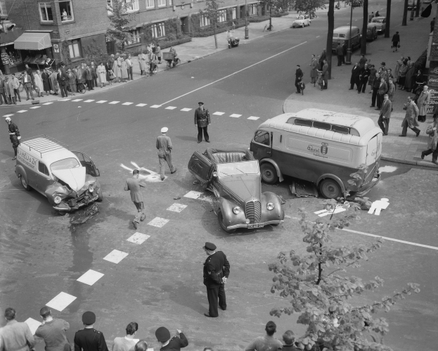 Eine Schwarz-Weiß-Szene eines Autounfalls an einer Straßenkreuzung mit mehreren Fahrzeugen, darunter ein Lieferwagen, einer Gruppe von Menschen drumherum und im Hintergrund Gebäude, Bäume und Masten.