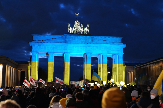 Eine Menschenmenge hält Fahnen und Plakate vor dem Brandenburger Tor in Berlin, Deutschland, wobei die Statuen und Säulen des Tores sowie ein bewölkter Himmel zu sehen sind; ein Protest-spezifisches Banner ist teilweise auf der rechten Seite sichtbar.
