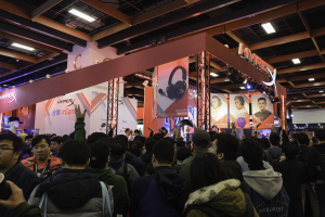 Eine große Menschenmenge versammelt sich vor einem Stand auf einer Gaming-Messe in China, einige tragen Taschen und einer hält einen Becher; Banner und Deckenbeleuchtung sind im Hintergrund zu sehen.