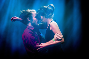 Zwei Menschen, die sich auf einer Bühne umarmen und küssen, vor minimalistischem Hintergrund.