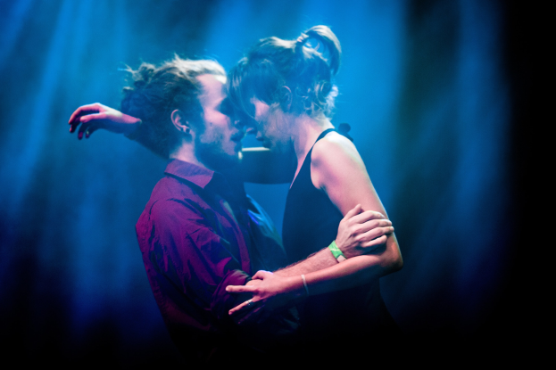 Zwei Menschen, die sich auf einer Bühne umarmen und küssen, vor minimalistischem Hintergrund.