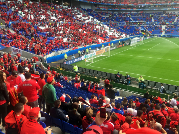 Eine große Menschenmenge in einem Stadion schaut einem Fußballspiel zu, mit Menschen auf Sitzen und Stehplätzen, Schildern, einem Torpfosten und Deckenbeleuchtung.