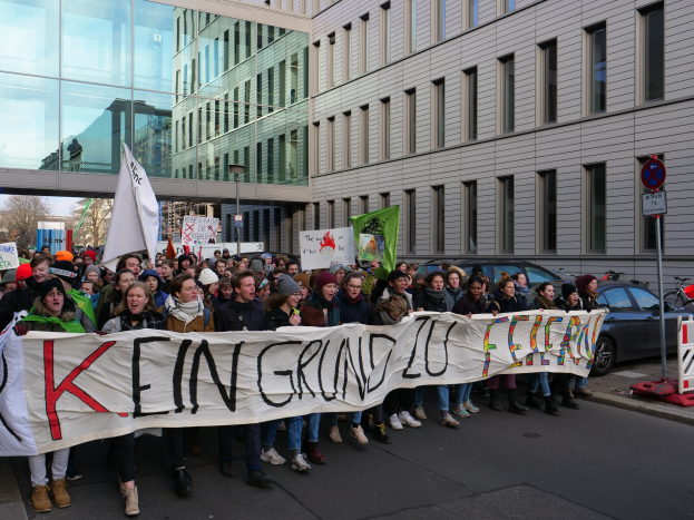 Eine Gruppe von Menschen, die auf einer Straße stehen und ein Banner mit der Aufschrift "Kein Grind Lo Feuer" halten, im Protest vor einem Gebäude, Bäumen, Polen, Schildern, Fahrzeugen und einem klaren blauen Himmel.