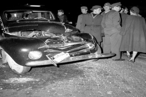 Ein Schwarz-Weiß-Bild eines verunglückten Autos am Straßenrand, umgeben von einer Gruppe besorgter Menschen, die auf die schweren Schäden an der Motorhaube und den zerbrochenen Fenstern starren.