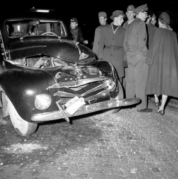 Ein Schwarz-Weiß-Bild eines verunglückten Autos am Straßenrand, umgeben von einer Gruppe besorgter Menschen, die auf die schweren Schäden an der Motorhaube und den zerbrochenen Fenstern starren.