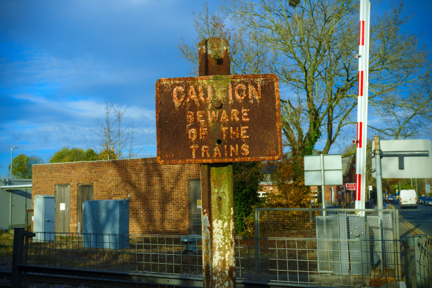 Achtung-Schild an einem Bahnübergang-Zaun neben Bäumen, Pfählen, einem Gebäude mit Fenstern, Containern, einem Straßenpfahl, Fahrzeugen auf einer Straße und einem bewölkten Himmel.