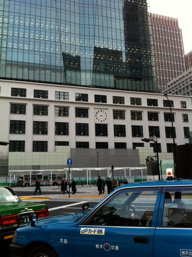 Eine Szene mit mehreren Autos, Holzplanken, einem Bus, Pfählen, Gebäuden und einigen Menschen, mit lesbarem Text auf einem der Autos.