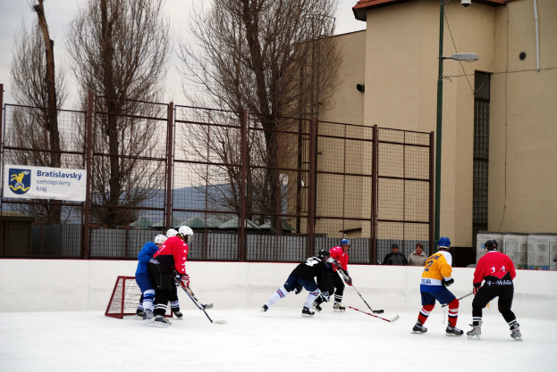 Personen beim Eishockeyspielen auf einem Eisstadion mit Gebäuden, Bäumen, Straßenlaterne, Namensschild und Zäunen im Hintergrund unter einem Himmel.