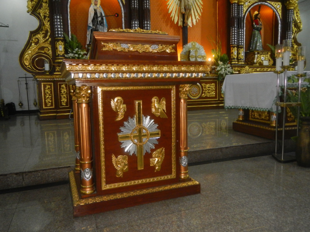Ein Altar in einer Kirche mit einem Kreuz, Statuen, Blumensträußen, Kerzen, einem tischdecke und anderen Gegenständen, mit Wänden, die mit Lichtern geschmückt sind.