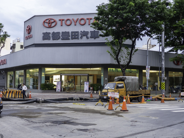 Eine Toyota-Händlerwerkstatt in Tokio, Japan, mit Fahrzeugen auf der Straße, Verkehrskegeln, Rohren, Menschen, Bäumen, Polen, Brettern und Gebäuden im Vordergrund und einem bewölkten Himmel im Hintergrund.