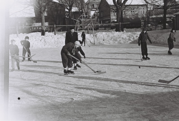 Eine Gruppe von Menschen, die Hockey auf einem Eisplatz umgeben von Schnee, Bäumen, Gebäuden mit Fenstern, Treppen mit Geländern und einem Lastwagen im Hintergrund spielen, dargestellt in Schwarz-Weiß.
