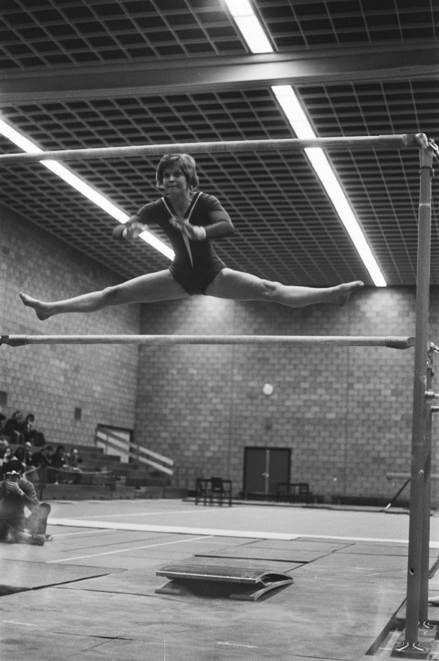 Eine Frau führt eine Gymnastikübung an den Stufenbarren in der Luft aus, mit ausgestreckten Armen und gebeugten Beinen, in einem Schwarz-Weiß-Bild eines Turnhallen mit Zuschauern auf der linken Seite und einer Tür im Hintergrund.