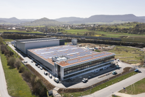 Luftaufnahme eines großen Gebäudes mit Solarmodulen auf dem Dach, umgeben von Fahrzeugen, Bäumen, Gras und Hügeln unter einem klaren blauen Himmel, identifiziert als Deutschlands erste solarbetriebene Fabrik.