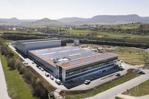 Luftaufnahme eines großen Gebäudes mit Solarmodulen auf dem Dach, umgeben von Fahrzeugen, Bäumen, Gras und Hügeln unter einem klaren blauen Himmel, identifiziert als Deutschlands erste solarbetriebene Fabrik.