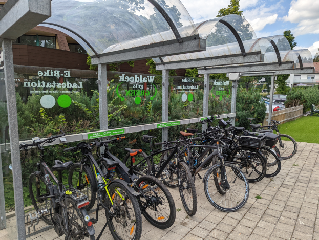 Eine Reihe von Fahrrädern, die vor einem Glasgebäude mit einem Schuppen geparkt sind, umgeben von Bäumen, Pflanzen und Fahrzeugen auf der Straße, mit einer bewölkten Himmel. Die Glaswände des Gebäudes zeigen Text, der auf ein E-Bike-Netzwerk in den Niederlanden hinweist.