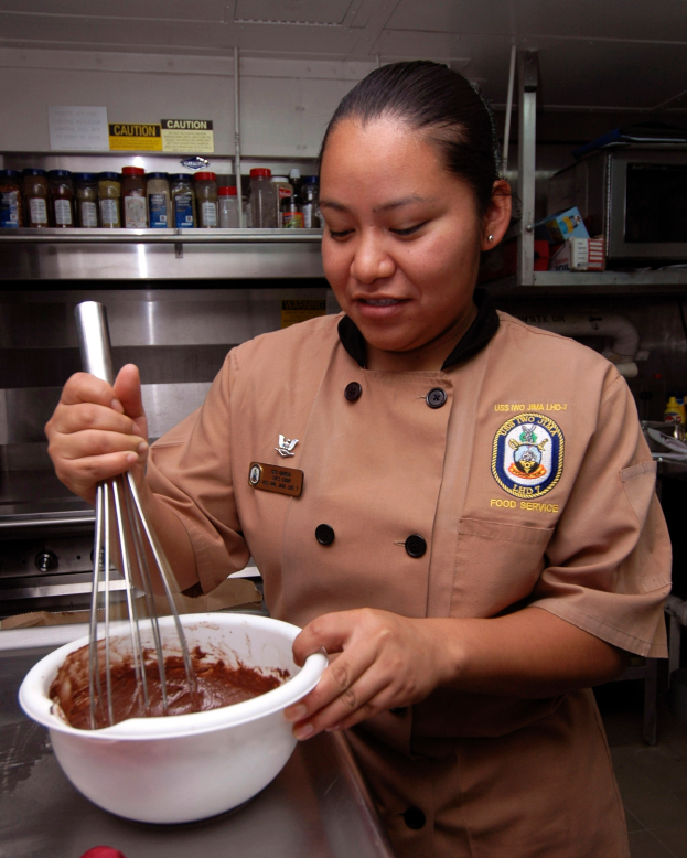 Eine Frau in Kochuniform steht in einer Küche und hält eine Schüssel und einen Schneebesen in der Hand, während verschiedene Gegenstände auf dem Tisch und Flaschen auf Gestellen im Hintergrund zu sehen sind.