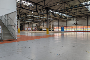 Ein großes industrielles Lagerhaus mit zahlreichen Fenstern, Metallstützen, einer Treppe mit Geländer, einem Schild, Deckenleuchten und zusätzlichen Stützen.