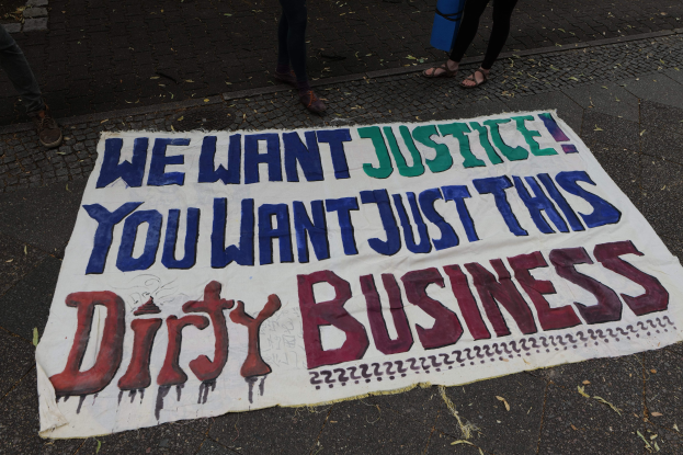 Banner auf einer Straße mit dem Text "Wir wollen Gerechtigkeit, du willst nur diesen schmutzigen Deal" und zwei Personen oben stehend, wobei eine Person ein blaues Objekt hält.