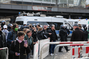 Eine Gruppe von Menschen steht vor Polizeifahrzeugen, einige tragen Helme und halten Telefone, mit Absperrungen im Vordergrund und einer Brücke mit Geländern und Gebäuden im Hintergrund, in Berlin, Deutschland.