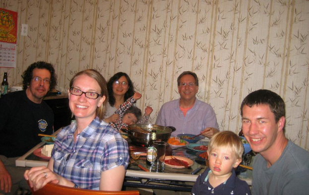 Ein Esstisch voller Gerichte und Essen in einem Wohnzimmer mit floralen Wanddesigns, umgeben von fünf strahlenden Menschen, mit einem Kalender und einer Flasche links daneben und einem Stuhl im Vordergrund.