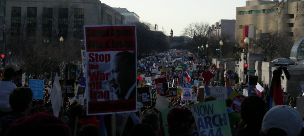 Eine Gruppe von Menschen protestiert auf einer Straße, hält Plakate, mit Gebäuden, Bäumen und Laternen im Hintergrund unter einem klaren Himmel.