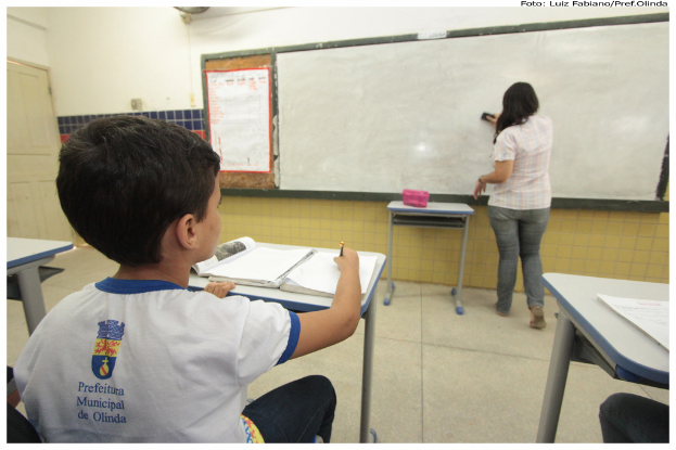 Ein Junge sitzt an einem Tisch und schreibt in ein Buch, während eine Frau die Tafel mit einem Putzlappen abwischt; zusätzliche Tische, eine Kiste und ein Plakat sind im Klassenzimmer zu sehen.