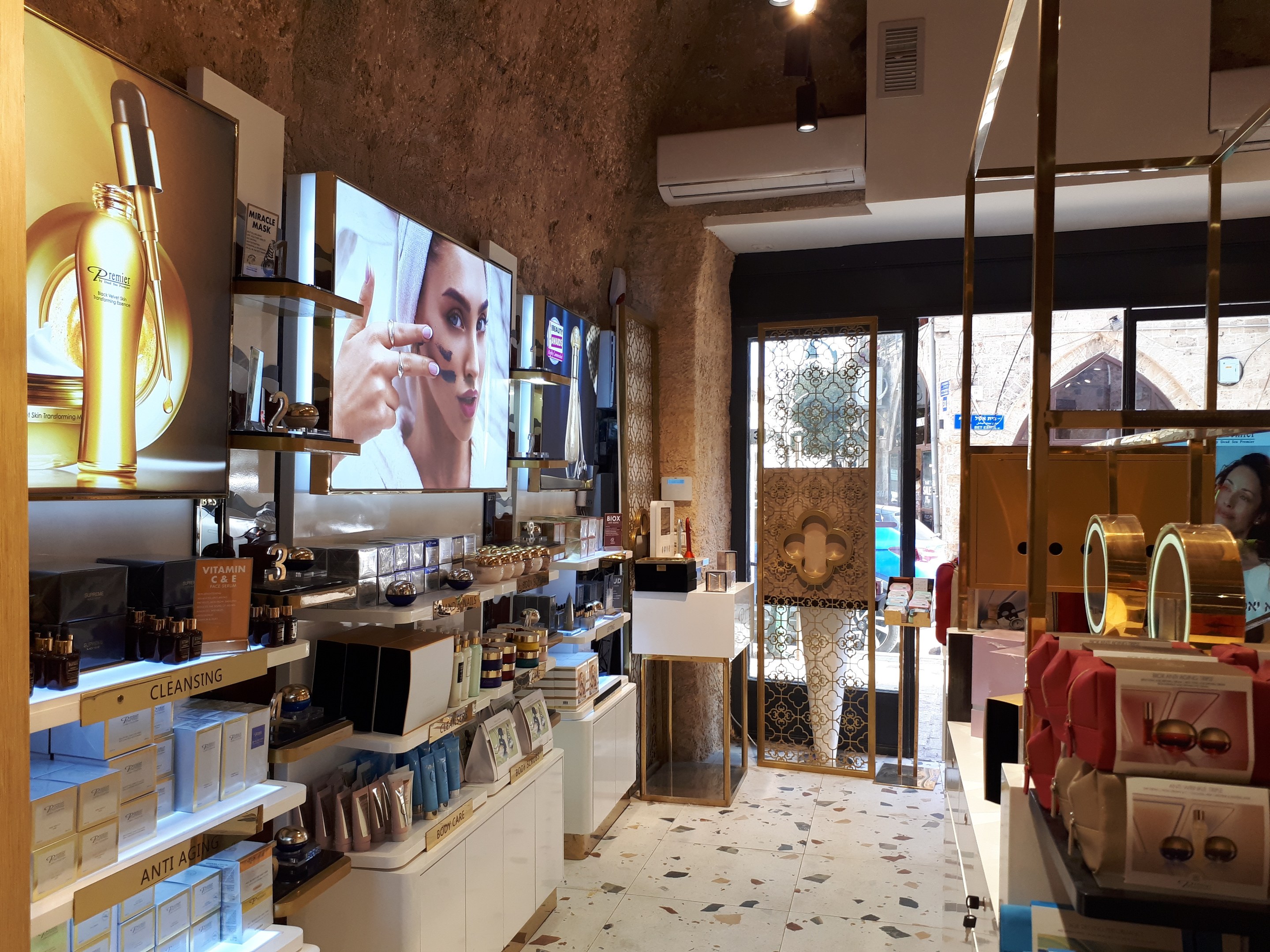 Innenansicht eines Kosmetikgeschäfts mit verschiedenen Produkten auf Gestellen, mit Wandbildschirmen, Deckenleuchten, einer Klimaanlage und Glastüren, die eine Person, ein Auto und ein Gebäude draußen zeigen.