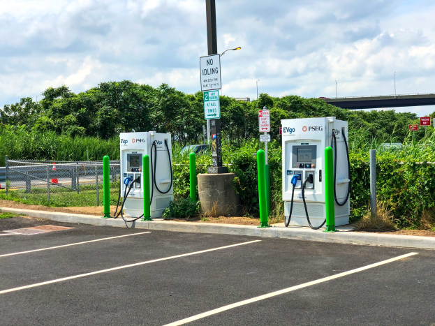 Elektrofahrzeug-Ladestation auf einem Parkplatz mit umgeben-der Infrastruktur, Vegetation und einer Brücke im Hintergrund unter einem bewölkten Himmel.
