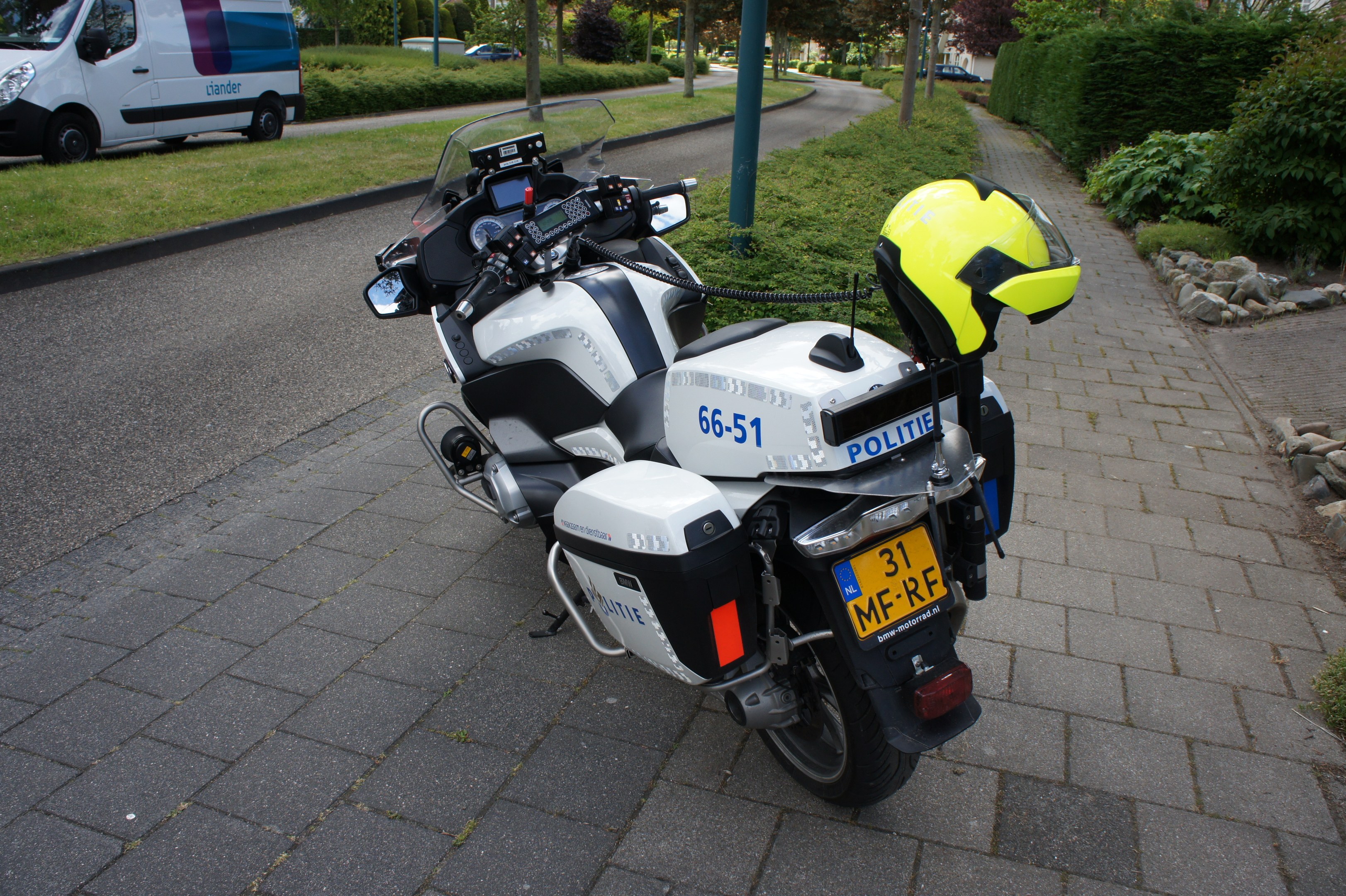 Ein Polizei-Motorrad am Straßenrand geparkt mit einem Helm darauf, umgeben von Bäumen, Polen, Gebäuden, Pflanzen, Gras, Steinen und anderen Fahrzeugen.