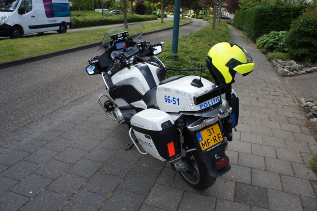 Ein Polizei-Motorrad am Straßenrand geparkt mit einem Helm darauf, umgeben von Bäumen, Polen, Gebäuden, Pflanzen, Gras, Steinen und anderen Fahrzeugen.