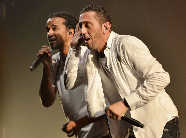Zwei Männer in legerer Kleidung stehen nebeneinander auf einer Bühne, jeder hält ein Mikrofon während einer Vorstellung, mit einem unscharfen Hintergrund.