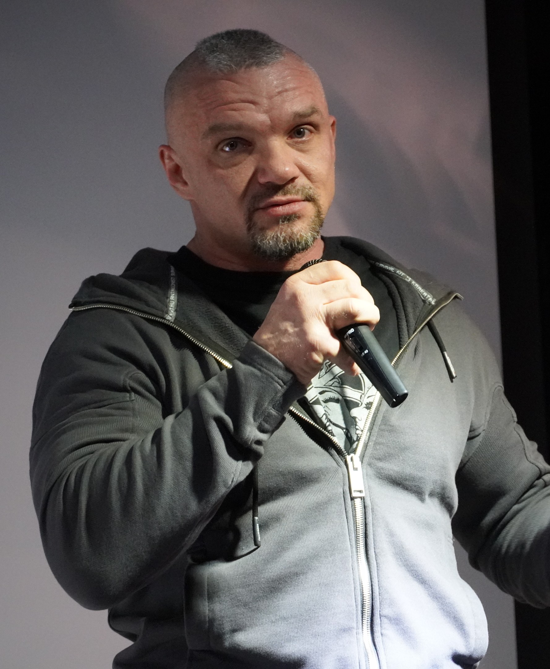 Ein Mann in einer schwarzen Kapuzenjacke und einem schwarzen Anzug mit einem weißen Hemd, der ein Mikrofon hält, steht selbstbewusst vor einer Wand und scheint ein Kom├Ądiant mit zurückgek├Ąmmtem Haar zu sein.