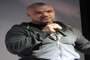 Ein Mann in einer schwarzen Kapuzenjacke und einem schwarzen Anzug mit einem weißen Hemd, der ein Mikrofon hält, steht selbstbewusst vor einer Wand und scheint ein Kom├Ądiant mit zurückgek├Ąmmtem Haar zu sein.