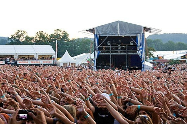 Menschenmenge auf einem Musikfestival mit erhobenen Händen, einige halten Handys, vor einer Bühne unter einem klaren blauen Himmel.