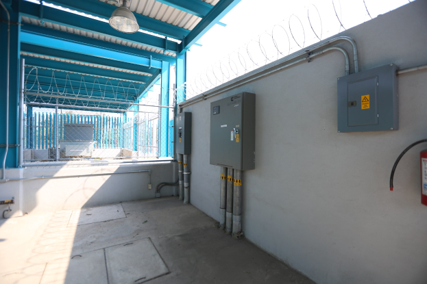 Ein Raum in einer elektrischen Umspannstation mit einem Feuerlöscher auf der rechten Seite, elektrischen Schaltkästen und Rohren an der Wand, einer Decke mit einer Lampe oben und einem Zaun im Hintergrund.
