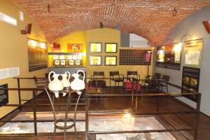 Ein Museumsraum voller Tische, Stühle, eines Geländers, Vasen, Texttafeln, Wandrahmen, Lampen und diverser Ausstellungsstücke.