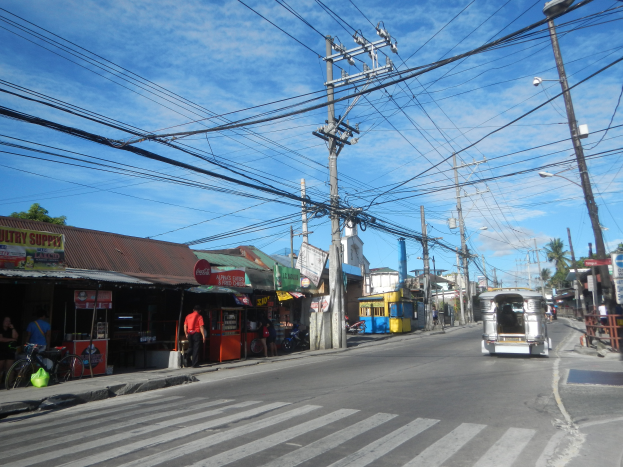 Eine Straße in einer Stadt mit elektrischen Masten und Kabeln, Fahrzeuge auf der Straße, Füßgänger auf dem Gehweg, abgestellte Fahrräder, Geschäfte mit Schildern, Bäume und ein bewölkter Himmel im Hintergrund.