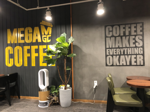 Ein Kaffeehaus-Interieur mit Tischen, Stühlen, einem "Mega Coffee"-Schild, einem Topf mit Pflanze, einem Bodenreinigungsgerät und Deckenleuchten.