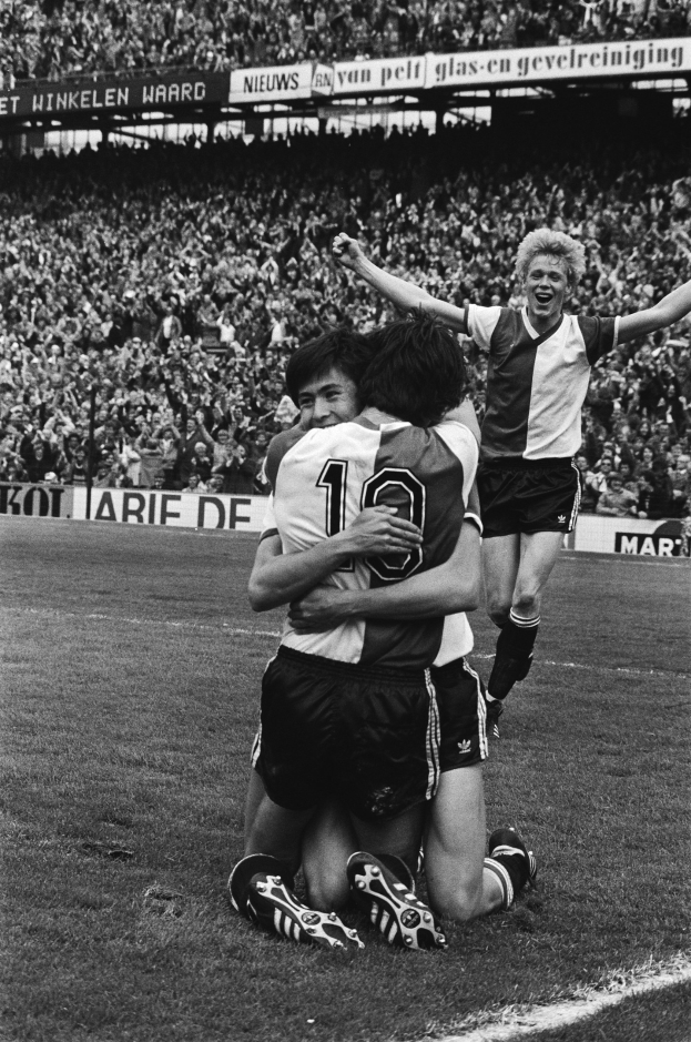 Drei Fußballspieler feiern ein Tor auf dem Boden, umgeben von einer jubelnden Menge und schwenkenden Fahnen in einem schwarz-weißen Stadion.