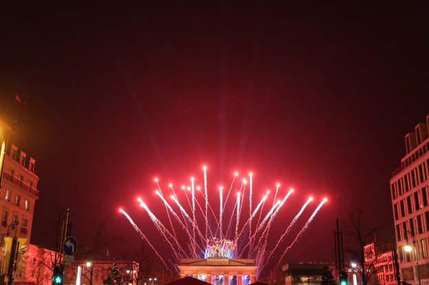 Eine belebte Straßenkreuzung in Berlin, Deutschland, mit dem Brandenburger Tor in der Mitte, beleuchtet von Gebäudelichtern, Bäumen, Pfosten, Verkehrsampeln, Schildern und Fahnen, mit Feuerwerk, das den Nachthimmel erhellt.