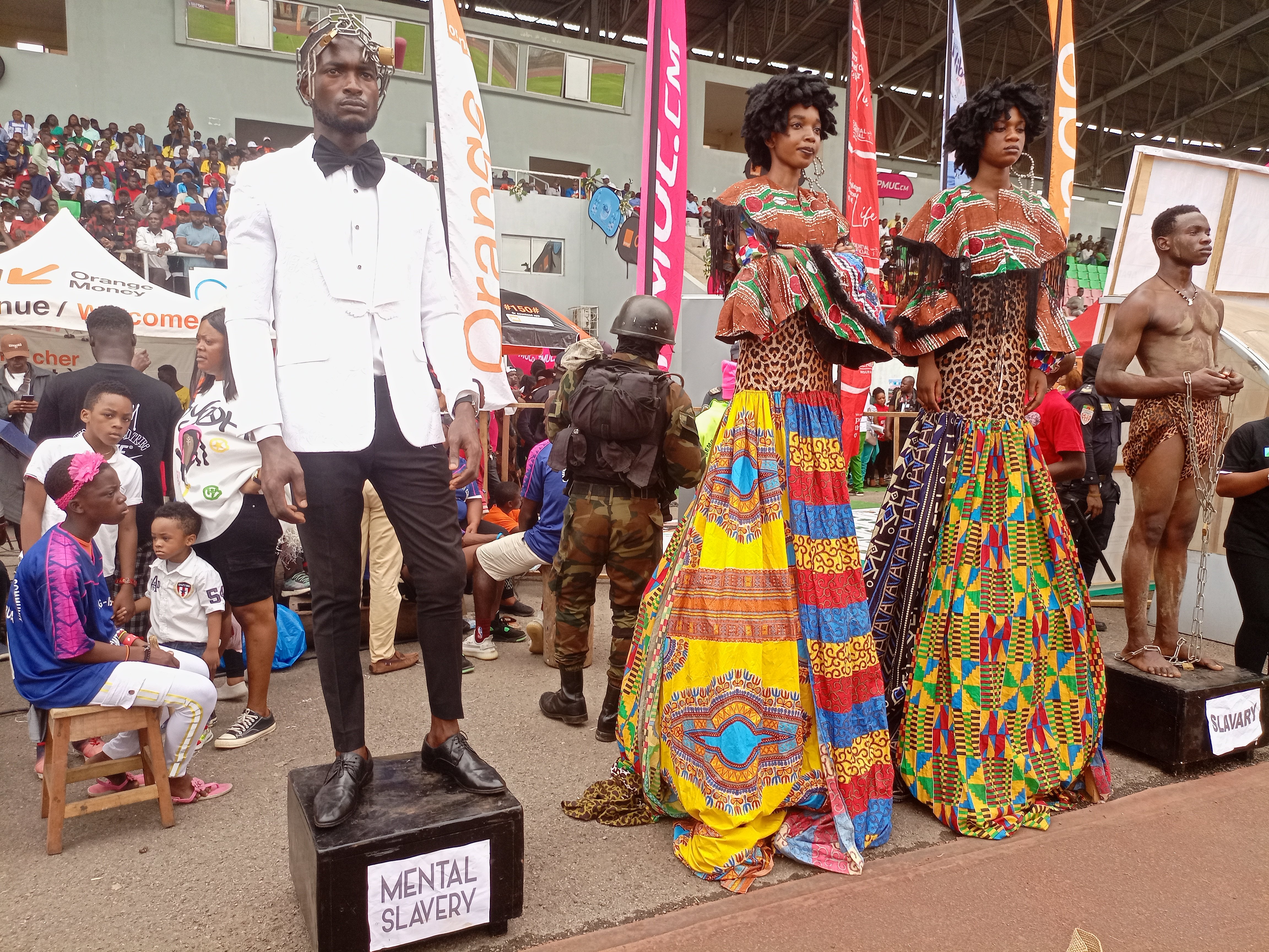Eine Gruppe von Menschen auf einer Bühne mit einer Schaufensterpuppe in einem weißen Anzug in der Mitte, andere auf Hockern links sitzend, eine Person mit einer Kamera rechts stehend und Banner, Flaggen und ein Gebäude im Hintergrund bei einer Fashion Show in Nigeria.