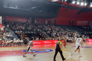 Eine Gruppe von Menschen, die Basketball in einer Turnhalle spielen, mit einem Basketballkorb links und Zuschauern im Hintergrund, unter hellen Stadionlichtern.