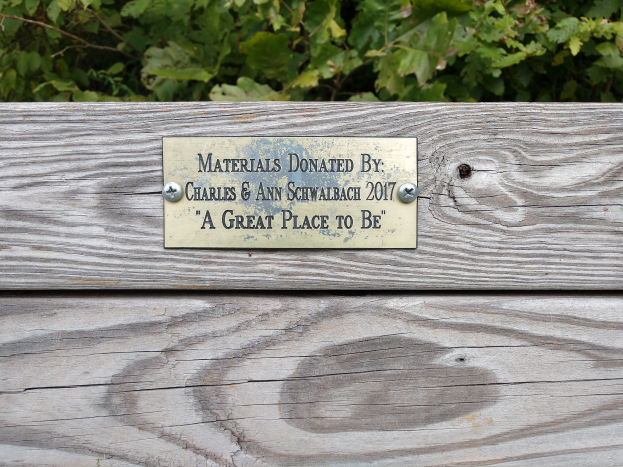 Eine hölzerne Bank mit einer Plakette, auf der "Materialien gespendet von Charles & Ann Schwabach 2017: Ein toller Ort" steht, umgeben von üppigen grünen Pflanzen.