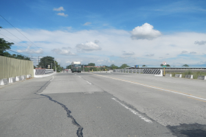 Eine Autobahn mit einem großen zentralen Riss, gesäumt von Fahrzeugen, einem Zaun, Bäumen, Strommasten mit Drähten, Schildern und einem bewölkten Himmel.