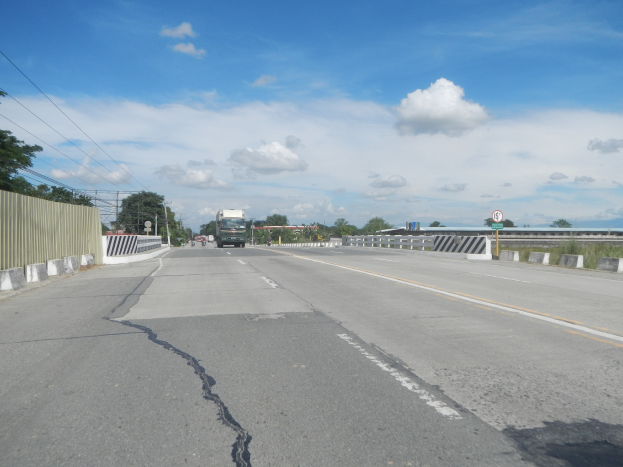 Eine Autobahn mit einem großen zentralen Riss, gesäumt von Fahrzeugen, einem Zaun, Bäumen, Strommasten mit Drähten, Schildern und einem bewölkten Himmel.